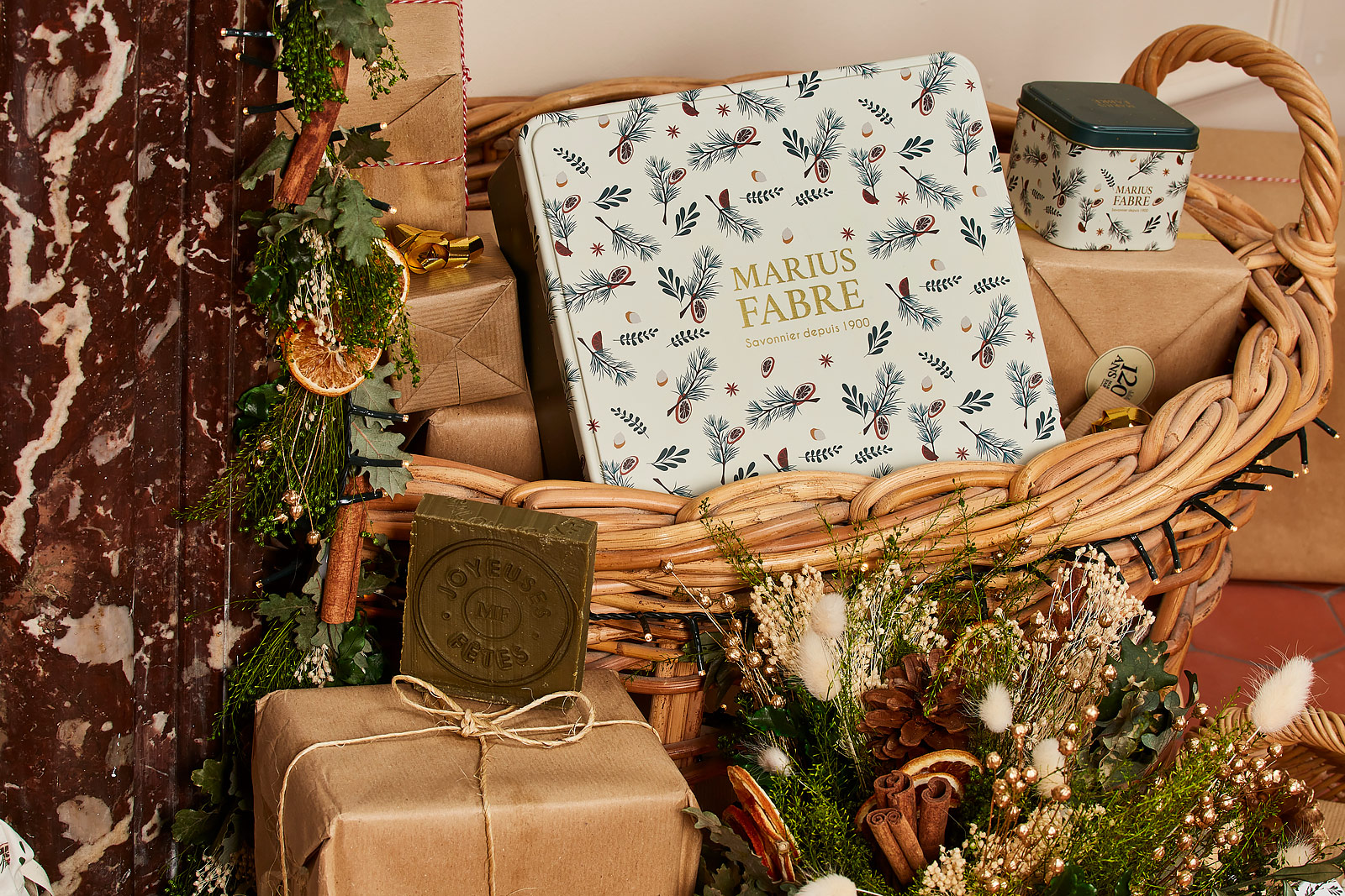 Ensemble de produits de la marque Marius Fabre mis en scène au pied d'une cheminée dans un panier en osier décoré d'un bouquet de fleurs séchées